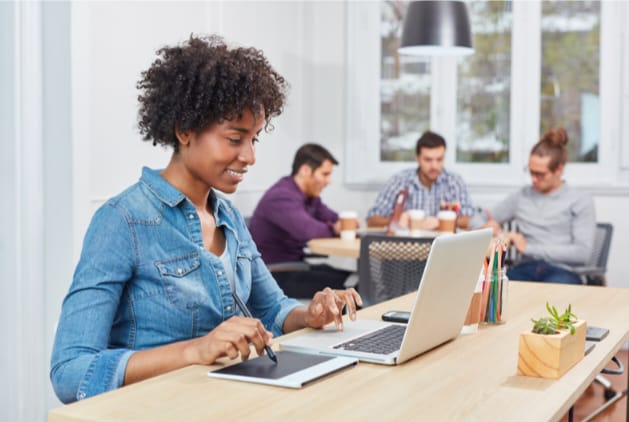 Mulher no computador pesquisando sobre benefício corporativo de terapia online e uma equipe reunida em uma mesa ao fundo.