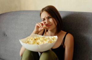 Mulher jovem comendo pipoca e assistindo dorama sobre saúde mental na TV.